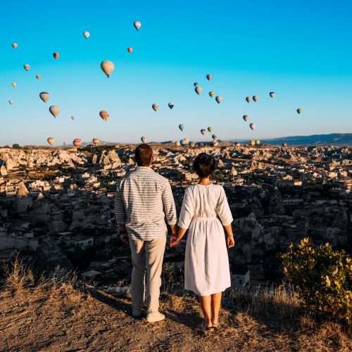 Hot air balloon flying over spectacular Cappadocia - couple watching hot air balloons at the hill of Cappadocia. Couple standing by each other in Turkey Cappadocia with hot air balloons at sunrise.